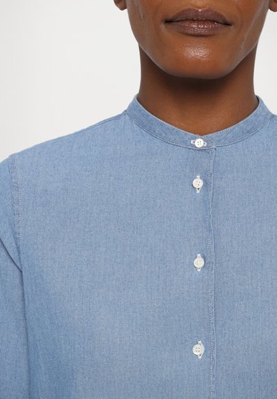 Lyseblå denimskjorte med mandarin-krave, udstyret med fem hvide knapper foran. Glat tekstur og let tætsiddende design.