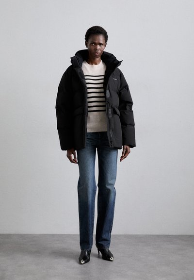Black puffer jacket with a high collar, front pockets, and a matte finish. Worn over a light striped sweater and blue jeans.