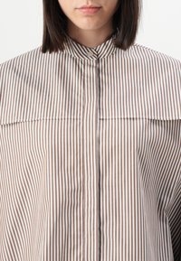 Camicia a strisce in cotone marrone e bianco. Presenta un colletto alto, una chiusura con bottoni e una vestibilità ampia e rilassata.
