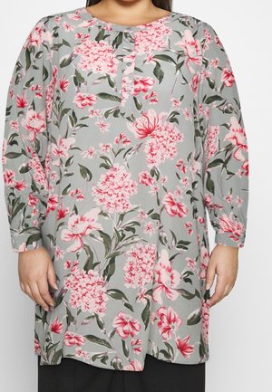 Femme portant une tunique grise à manches longues avec des motifs floraux roses et un col boutonné, debout devant un fond uni.