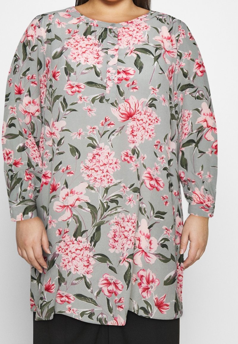 Femme portant une tunique grise à manches longues avec des motifs floraux roses et un col boutonné, debout devant un fond uni.