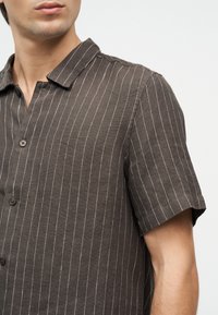 Homme portant une chemise à manches courtes foncée à boutons avec de fines rayures blanches verticales et un col ouvert, sur un fond clair uni.