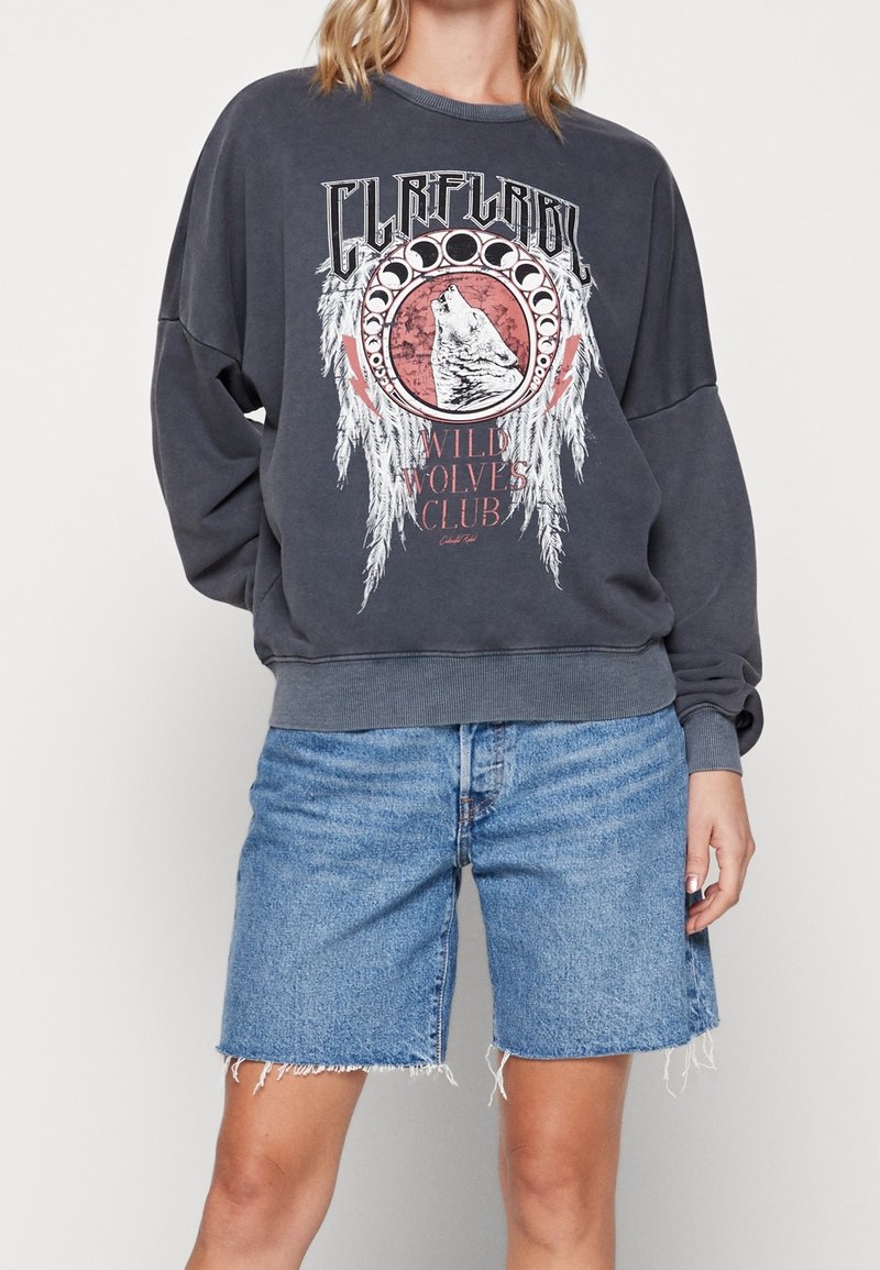 Femme portant un sweat-shirt gris foncé avec un motif de loup et des phases de lune, texte "Wild Wolves Club", associé à un short en denim bleu clair.
