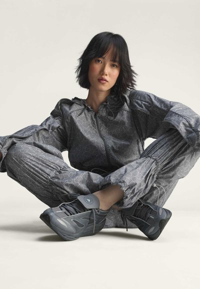 Woman with short black hair sits cross-legged wearing matching gray floral-patterned jacket and pants with gray athletic shoes against neutral backdrop.