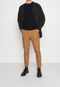 Musta neuletakki tummansinisen neulepuseron päällä, yhdistetty vaaleanruskeisiin kapealahkeisiin housuihin ja mustiin nauhakenkiin. Sileä kangas, rento istuvuus ja kontrastivärit.