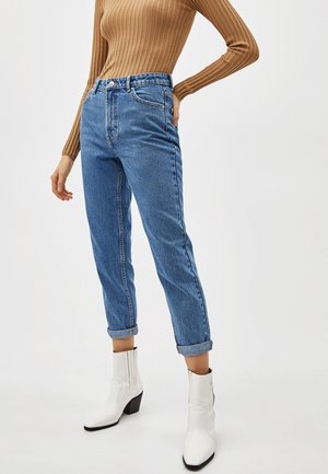 Osoba ubrana w podwinięte niebieskie dżinsy, brązowy prążkowany top z długim rękawem i białe botki za kostkę, stoi z jedną ręką na biodrze.