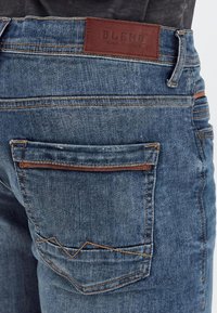 Poche arrière d’un jean en denim bleu avec une garniture en cuir marron et une étiquette de marque en cuir marron portant l’inscription "BLEND Global Jeansmaker" sur la taille.