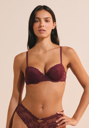 Giovane donna con lunghi capelli scuri che indossa un reggiseno in pizzo bordeaux e biancheria intima in pizzo coordinata, in piedi davanti a uno sfondo beige.