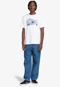 White cotton t-shirt with blue graphic design featuring a bus and "PARIS 1979" text. Paired with loose-fitting blue denim jeans and black sneakers.