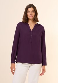 Blouse violette à manches longues avec une patte de boutonnage, un design minimaliste et une texture douce, associée à un pantalon blanc.