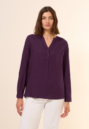 Blusa lunga viola a maniche lunghe con pattina bottonata, design minimalista e texture morbida, abbinata a pantaloni bianchi.
