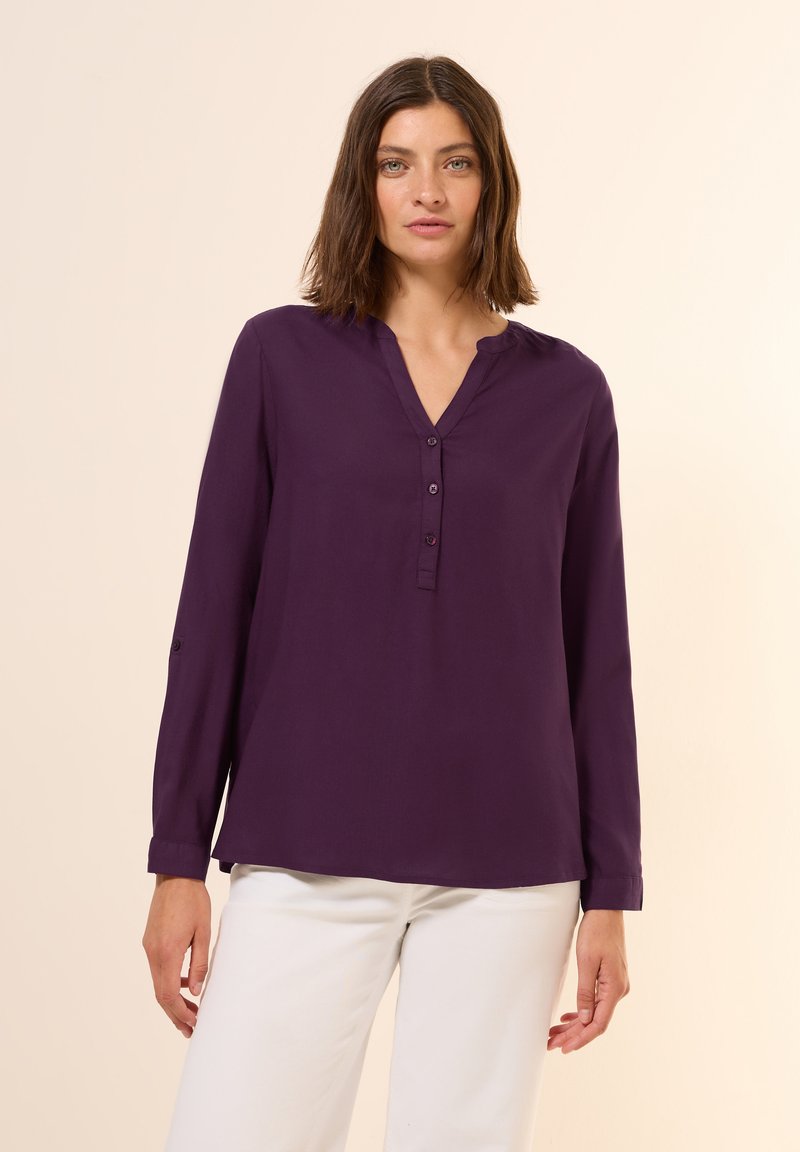 Blouse violette à manches longues avec une patte de boutonnage, un design minimaliste et une texture douce, associée à un pantalon blanc.