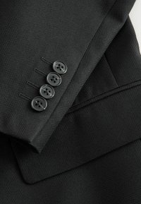 Manche de veste de costume texturée noire avec quatre boutons noirs assortis soigneusement alignés près du poignet et une partie du revers visible.