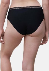 Frau in schwarzer, hoch geschnittener Unterwäsche mit Spitze, von hinten vor einem schlichten weißen Hintergrund gezeigt.