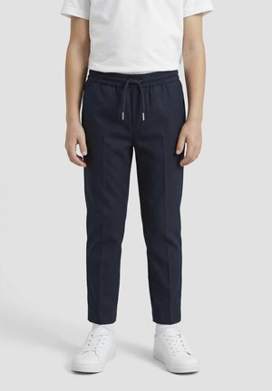 Niño con pantalones deportivos azul marino con cintura de cordón, calcetines blancos, zapatillas blancas y una camiseta blanca lisa de manga corta.