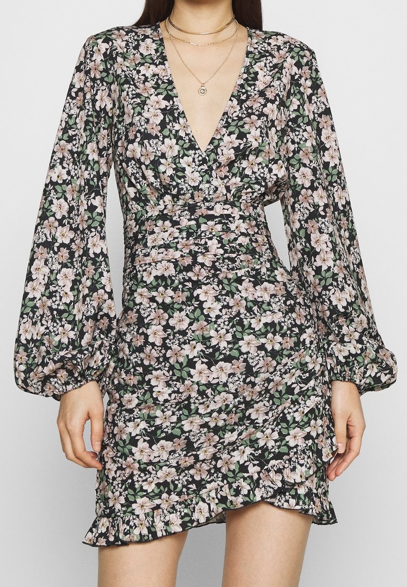 Robe noire à fleurs avec de longues manches bouffantes, un profond décolleté en V et une taille froncée. Elle présente de petites fleurs roses et blanches ainsi qu'un ourlet à volants.