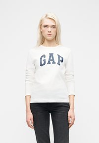 Jeune femme aux cheveux blonds portant un t-shirt blanc à manches longues avec le logo "GAP" et un jean noir, debout devant un fond blanc uni.