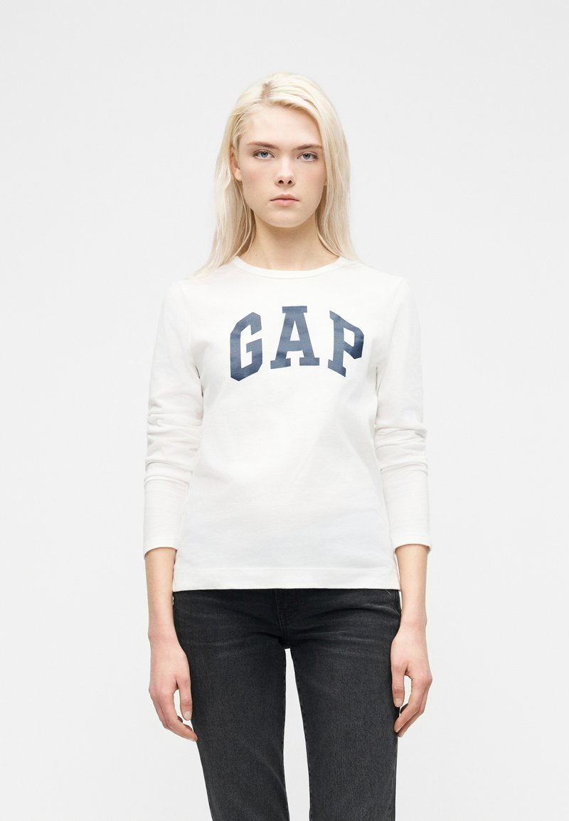 Jeune femme aux cheveux blonds portant un t-shirt blanc à manches longues avec le logo "GAP" et un jean noir, debout devant un fond blanc uni.