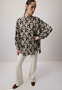 Bluză cu model negru și crem, cu guler înalt și mâneci tip balon, purtată cu pantaloni largi albi și pantofi slip-on maro.