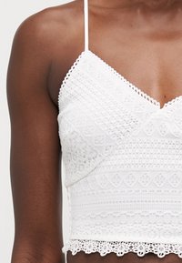Valkoinen pitsinen bralette, jossa on monimutkaisia kuvioita, V-mallinen kaula-aukko, säädettävät spagettiolkaimet ja kaartuva helma, joka korostaa rakennepintaa.