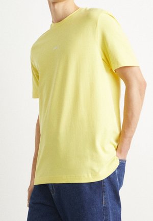 Homme portant un t-shirt jaune uni à manches courtes et un jean bleu, avec une main dans la poche, sur un fond blanc uni.