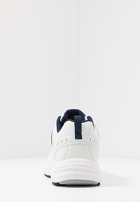 Hvit sneaker med marineblå aksenter, som har en teksturert mesh-tunge, sømdetaljer og et trekkpunkt på hælen. Gummisåle synlig.