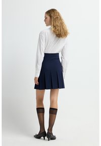 Falda plisada azul marina combinada con una camisa blanca de manga larga a rayas. La modelo lleva puestas medias altas negras y zapatos de tacón negros.