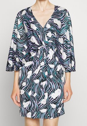 Vrouw draagt een jurk tot op de knie met een voorpand in een gedraaide knoop, voorzien van een multikleuren abstract patroon in zwart, wit, paars en teal.