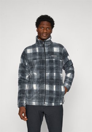 Man wearing a gray and white plaid zip-up fleece jacket with hands in pockets, standing against a plain light background.