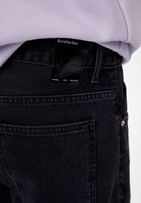 Svarta denimjeans med en stilren design, som har en glansig svart etikett vid midjan, synlig sömnad och en enda bakficka.