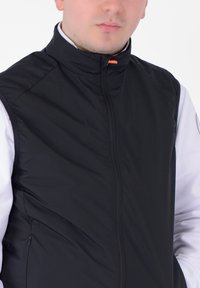 Uomo che indossa un gilet nero con cerniera sopra una camicia bianca a maniche lunghe, con le mani nelle tasche del gilet, su uno sfondo semplice.