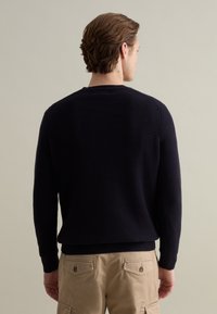 Maglione lavorato a maglia blu navy con un motivo testurizzato, polsini a coste e scollatura rotonda, visto da dietro. Vestibilità casual, indossato con pantaloni beige.