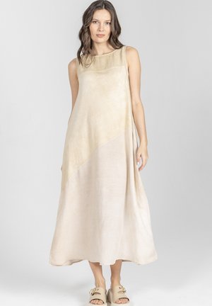 Kobieta w bezrękawnej maxi sukience w beżowo-kremowe pasy, z szerokimi beżowymi sandałami, stojąca na jednolitym jasnym tle.