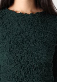 Dark green textured top with a wavy neckline. The fabric appears crinkled and layered, creating a distinct surface pattern.