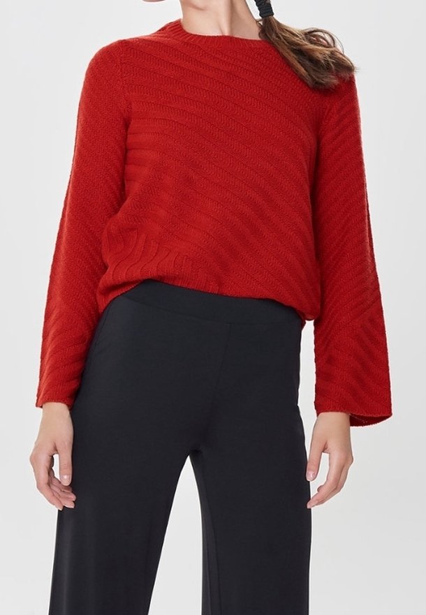 Femme portant un pull rouge texturé à manches larges et un pantalon noir ample, debout devant un fond neutre.