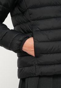 Svart pufferjakke med quiltet design, synlig sidelomme og elastiske mansjetter. Materialet ser glansfullt ut og har en jevn tekstur.