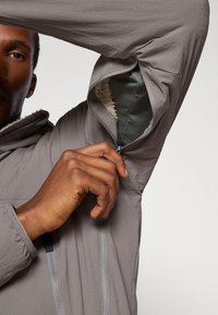 Veste grise pour hommes avec une poche zippée sur la manche, dotée d'une doublure intérieure douce. Texture lisse, coupe athlétique et design minimal.