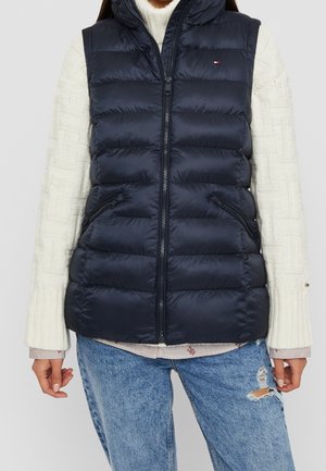 Frau trägt eine marineblaue gesteppte Weste über einem weißen Strickpullover und zerrissenen blauen Jeans, steht mit entspannten Armen seitlich am Körper.