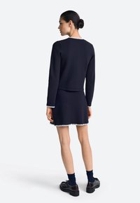 Tumesinine kahest osast koosnev komplekt, mis sisaldab pika varrukaga ülaosa ja sobivat seelikut. Mõlemal on valge laineline ääris ja tekstuurse pinnaga viimistlus. Mustad kingad.