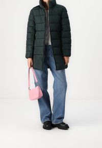 Persona vistiendo una chaqueta de plumas verde oscura, camiseta gris claro, jeans azules, zapatos negros, sosteniendo un pequeño bolso rosa por su correa con la mano izquierda.