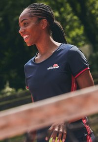 Camisa deportiva navy con acentos rojos y un logo, diseño de cuello redondo, mangas cortas y textura suave, llevada por una mujer que sostiene una pelota de tenis.