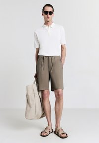 Beige Polo-Shirt, khakifarbene Shorts und flache Sandalen. Modell hält eine hellbeige Tragetasche. Glatte Texturen, minimalistisches Design.