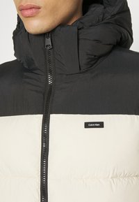 Homme portant une doudoune Calvin Klein noir et blanc, zippée jusqu'au cou avec un col haut matelassé et une capuche.