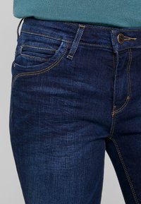 Jeans de mezclilla azul oscuro con una textura suave, acentos de costura dorada, bolsillos delanteros y un diseño clásico de cierre de botón.
