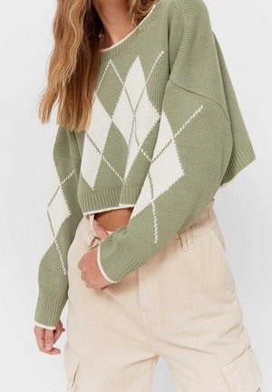 Groene gebreide trui met een wit ruitpatroon, cropped ontwerp, wijde mouwen en ribboorden, gecombineerd met lichtbeige broeken.