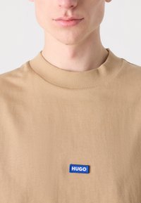 Tan kulatý výstřih tričko vyrobené z měkké tkaniny, na přední straně s modrým pravoúhlým logem "HUGO". Hladká textura a žebrovaný límec.