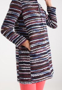 Abrigo tejido a rayas en navy, burdeos y blanco, con mangas largas, corte recto y bolsillos frontales. Llevado sobre pantalones rosas.
