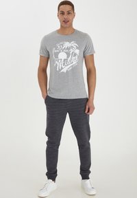 Camiseta de algodón gris con estampado gráfico blanco que presenta palmeras y un sol. Combinada con pantalones de chándal gris oscuro y zapatillas blancas.