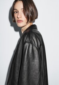 Černá kožená bunda s volným střihem, širokým límcem a viditelnými švy. Subjekt má krátké rovné vlasy a neutrální výraz.