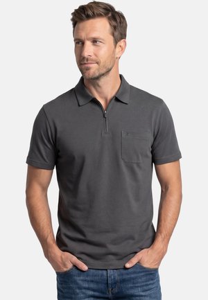 Mand med kort brunt hår iført en mørkegrå kortærmet poloshirt med lynlås foran og brystlomme, hænderne i lommene på jeans.